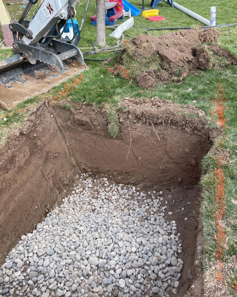 Trench in the grass with washed gravel ready for French drain installation.